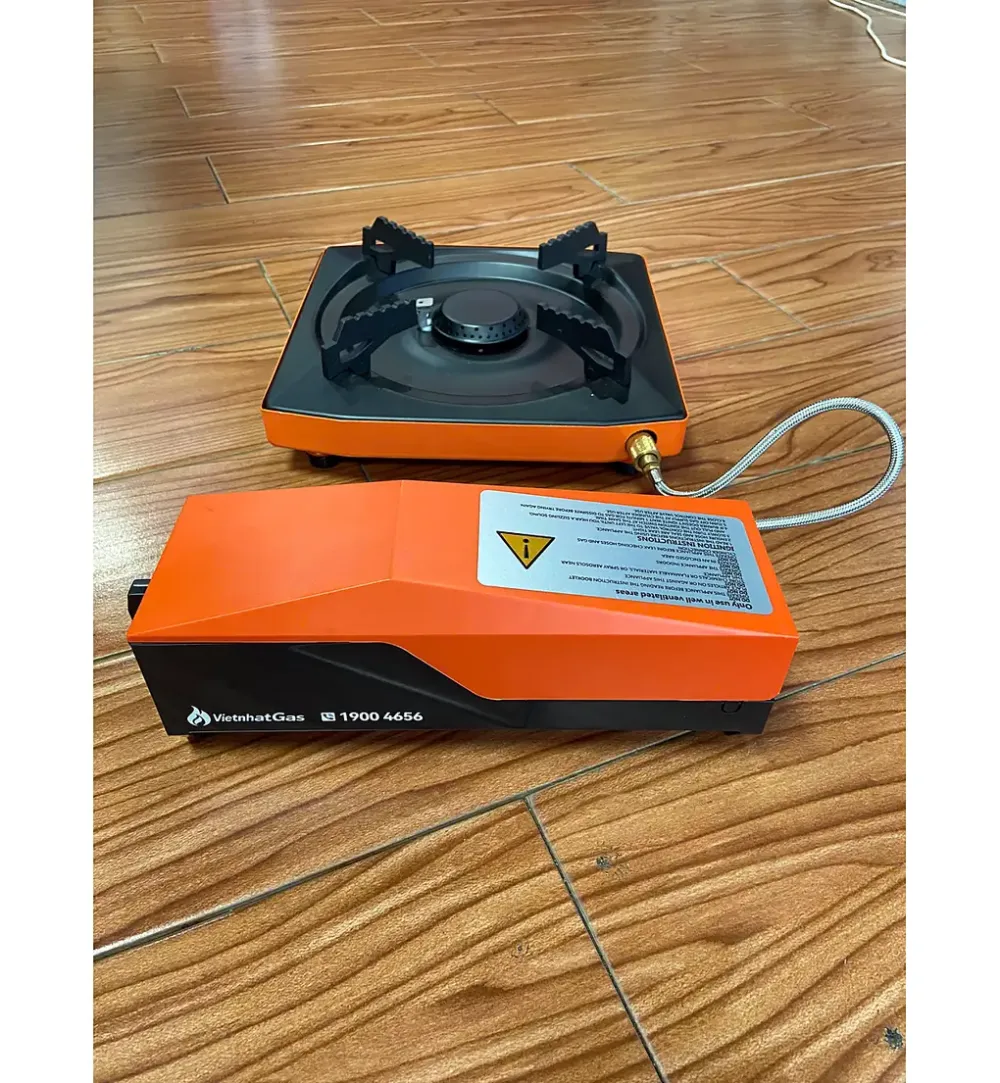 Bếp gas mini mặt đen nối ống dẫn với hộp gas rời màu cam trên sàn gỗ